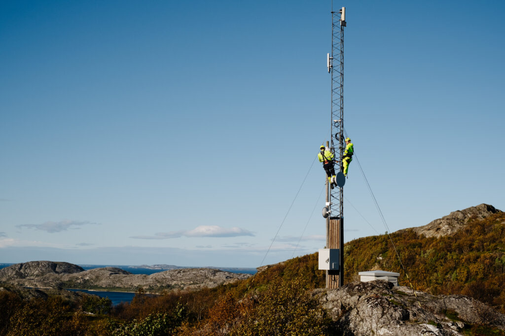 To montører står høyt oppe i en telemast på Keiservarden i Bodø. Vi ser Bodølandskapet i bakgrunnen.